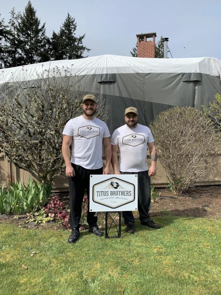 Two brothers standing together, smiling behind a company sign, wearing Titus Brothers hats and shirts during their first year of business.