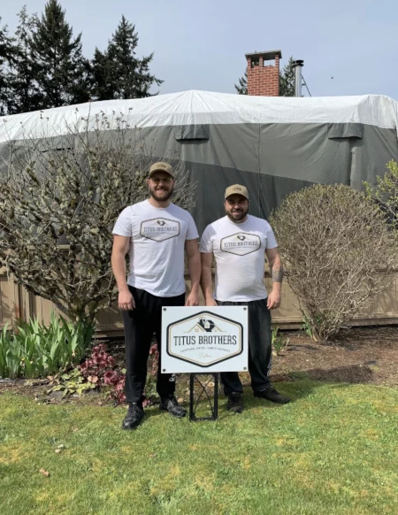 Two brothers standing together, smiling behind a company sign, wearing Titus Brothers hats and shirts during their first year of business.